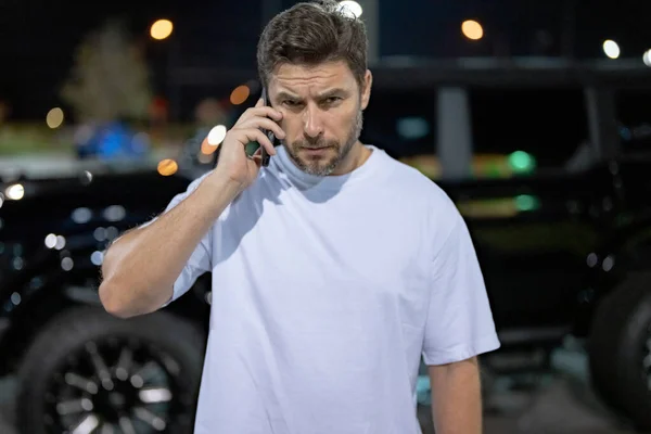 Gangster call phone. Angry man chatting on phone near car on night urban street. Dangerous aggressive man with phone. Criminal city. Danger american district. Aggressive angry man talking on phone