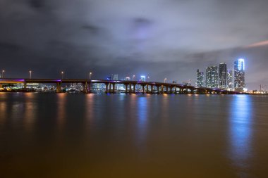 Günbatımında Miami manzarası, ABD. Alacakaranlıkta Miami City gökdelenleri ve deniz üzerindeki yansımalı köprü ile gökyüzü manzarası