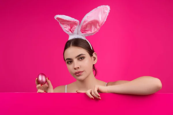 Young woman wearing bunny ears and peeking over a banner board. Beautiful young woman with bunny ears and blank poster on studio color background. Banner, copy space. Blank poster