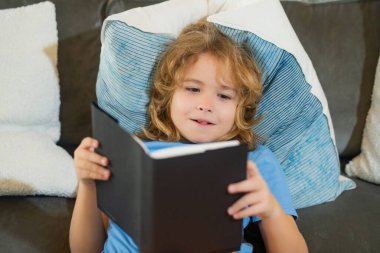 Hayali çocuk. Çocuk oturma odasında kitap okuyor. Çocuklar kitap okur. Küçük çocuk güneşli oturma odasında kanepede oturmuş masal kitabındaki resimleri izliyor.