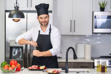 Mutfakta yemek pişiren yakışıklı bir adam. Mutfakta sebze ve etle yemek pişiren olgun bir adam. Kafkasyalı önlüklü adam ve aşçı şapkalı aşçı mutfakta lezzetli bir öğle yemeği pişiriyor. Akşam yemeği için sağlıklı yemek.