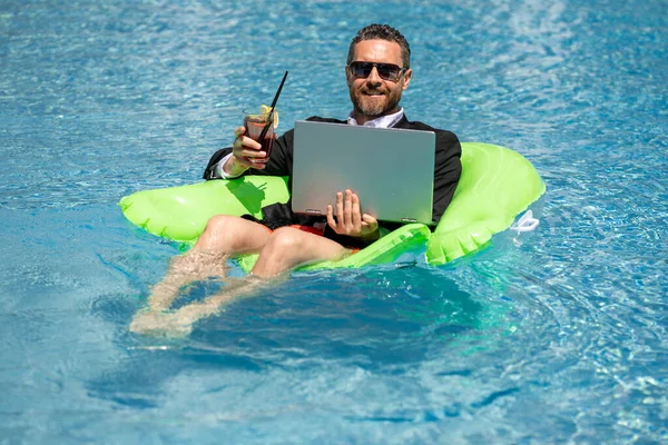 Freelance work, distance online work, e-working. Summer business. Business man in suit drink summer cocktail and using laptop in pool. Businessman dreams on summer business in swimming pool water