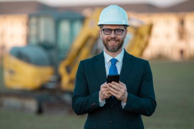 Başarılı bir inşaat işletmesi sahibi. Kazıcının yanındaki kask ve takım elbiseli inşaat işçisi. İnşaat alanının önünde kendine güvenen bir inşaat sahibi. İnşaat mühendisi.