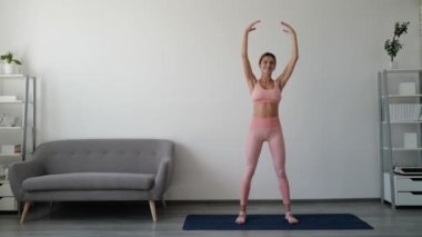 Tüm vücudunu evde spor yapan genç bir kadın çalıştırıyor. Fitness, spor, eğitim ve yaşam tarzı konsepti. Spor kıyafetli gülümseyen kadın paspasın üzerinde geriniyor.