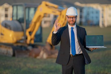 İnşaat mühendisi inşaat alanında çalışıyor. Olgun mühendis. İnşaat alanında takım elbiseli ve kasklı bir adam var. Orta yaşlı inşaat mühendisi kazıcının yanında duruyor.