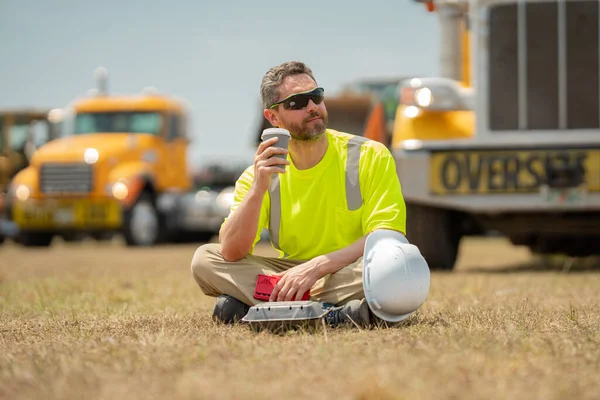 Worker taking break from work drinking coffee and resting sitting on grass. Builder worker relaxed after work on coffee break. Worker break relax time. Worker on lunch break. Truck driver