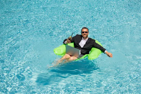 Successful business man floating in suit in pool water. Summer vacations and travel concept. Funny business man drink summer cocktail and using laptop in swimming pool. Summer business concept