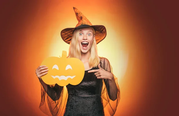 Beautiful surprised woman in witches hat and costume - showing products. Halloween Woman portrait. Beauty Woman posing with Pumpkin and Halloween hat. Helloween people concept