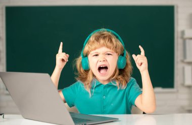 Kulaklıklı kızgın çocuk masada oturur, okulda dizüstü bilgisayarda çalışır, klever çocuk internet derslerini kullanarak öğrenirken kulaklık takar. Heyecanlı çocuk duyguları.