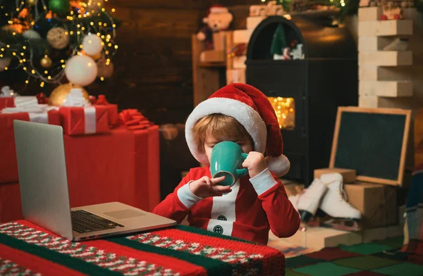小孩穿着圣诞老人的衣服坐在笔记本电脑旁 喝着热的圣诞饮料 固定背景 圣诞节的时候小男孩 圣诞老人 — 图库照片