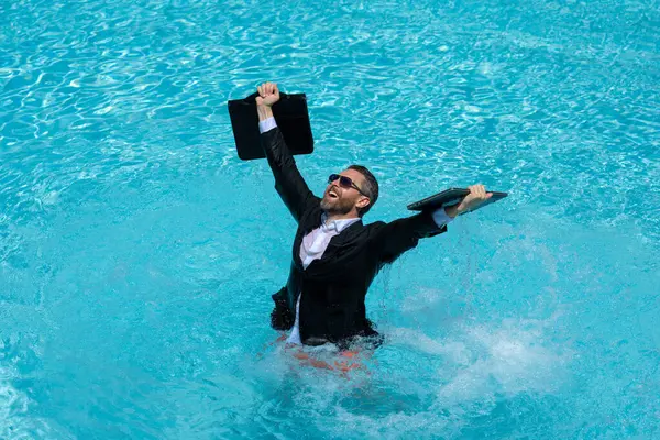 Funny business man in business suit with laptop standing in the water in the pool. Remote work. Crazy freelancer. Business and summer. Summer leisure weekend and remote freelance work. Crazy business