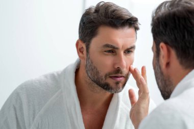 Bin yıllık İspanyol adam aynaya, yüz derisine ve sakalına bakıyor. Erkek güzellik ürünleri. Cilt bakımı, spa. Güzel bir adamın güzel portresi. Spa modeli, nemlendirici krem