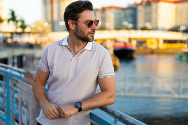 Şehir merkezinde sıradan, yakışıklı bir manken. İyi giyimli, gözlüklü ve polo pozu veren bir adam. Erkek model. Şehirde şık bir adamın portresi. Açık hava moda portresi