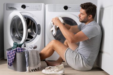 Çamaşır makinesinin yanında kirli giysileri olan seksi bir adamın portresi. Yakışıklı adam çamaşır makinesinin önünde oturur. Kirli çamaşırlar. Adam elbiseleri temizliyor. Yalnız bir adam için ev işi. Ev çamaşırları