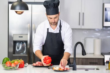 Önlüklü adam ve aşçı şapkalı adam mutfakta yemek pişiriyor. Mutfakta sağlıklı yemek pişiren yakışıklı adam. Adam mutfakta yemek pişiriyor. Taze yemek malzemeleriyle ev menüsü. Modern mutfak iç mimarisi