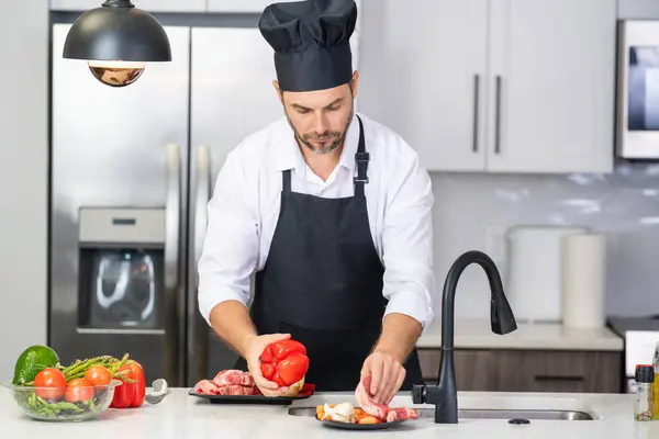 Man in apron and chef hat cooking food in kitchen. Handsome man cooking healthy food in kitchen. Guy cooking dinner food in kitchen. Home menu with fresh food ingredients. Modern kitchen interior