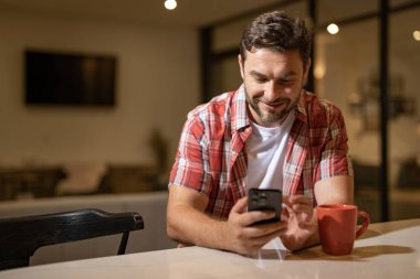 Hafta sonu evde oturan cep telefonu olan yakışıklı bir adam. Amerikalı bir adam oturma odasındaki koltukta otururken telefon kullanıyor. Akıllı telefonlu rahatlama metni. Telefon et.