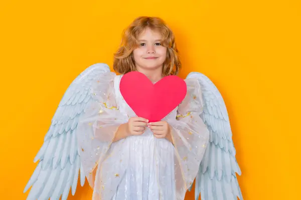 ペーパーハート 天使の翼を持つ子供は赤いハートを保持しています ハートシンボル愛とバレンタインデー バレンタインデー 黄色いスタジオの背景に天使の翼を持つブロンドかわいい子供 ハッピーエンジェル チャイルド — ストック写真