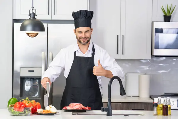 Man in apron and chef hat cooking food in kitchen. Handsome man cooking healthy food in kitchen. Guy cooking dinner food in kitchen. Home menu with fresh food ingredients. Modern kitchen interior