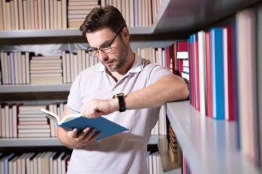 Okul kütüphanesinde orta yaşlı bir öğretmen kitap okuyor. Öğretmenler günü. Üniversite sınavı öğretmenleri. Üniversite öğretmeni. Öğretmek ve öğrenmek. Sınıfta kitabı olan bir öğretmen portresi.