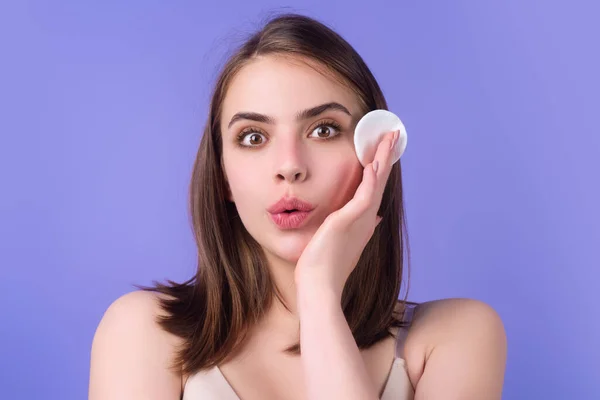 Beautiful young woman with cotton pad, applying lotion on face, removing makeup. Skin care. Studio portrait of girl cleaning her face with cotton pad, isolated background. Face toner and cotton pad
