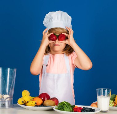 Aşçı çocuk sağlıklı yemek için taze sebze yaparken çilek tutuyor. Gri arka planda izole edilmiş küçük çocuk portresi. Aşçı çocuk. Yemek pişirme süreci