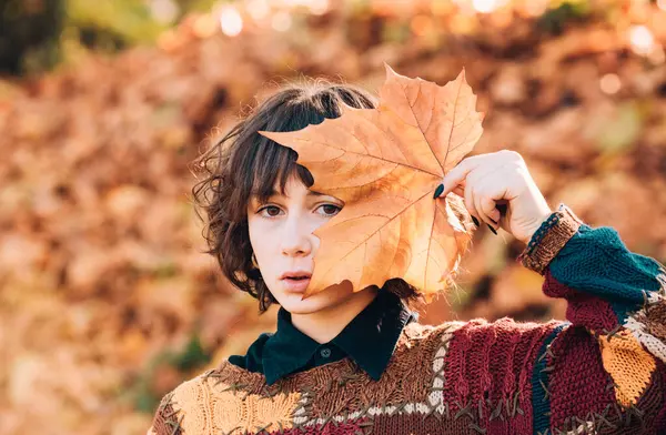 Beautiful girl with no make up. Attractive student girl wearing stylish sweater. Autumnal nature atmosphere. Close up portrait of pretty brunette hiding her face behind yellow leaf