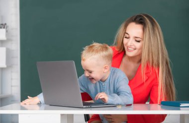 Anne ve oğlu birlikte bilgisayar laptopu kullanıyorlar. Küçük okul çocuğu annesiyle dizüstü bilgisayar kullanıyor. Öğretmenler okulda öğrencilere yardım ediyor.