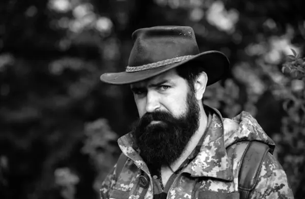 Portrait of adventure serious man extreme explorer. Man hunter in camouflage outdoor. Brutal hunter, bearded man in the wild forest in the autumn. Portrait man in cowboy hat