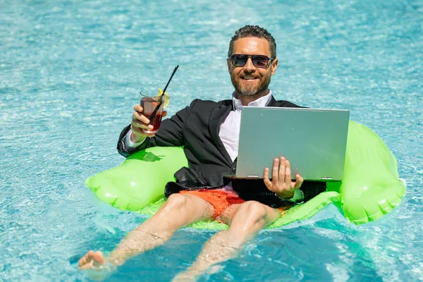 Successful business man floating in suit in pool water. Summer vacations and travel concept. Funny business man drink summer cocktail and using laptop in swimming pool. Summer business concept