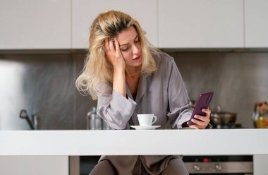 Mutfakta cep telefonuyla konuşan üzgün bir kadın. Sabahları ev hanımı modundayım. Sosyal medya kavramı