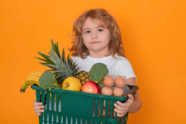 İndirim ve indirim. Sevimli çocuk alışveriş arabası ağzına kadar yiyecek dolu. Çocuk alışveriş sepetini sarı arka planda tutuyor. Mağaza, alışveriş, satış ve indirimler