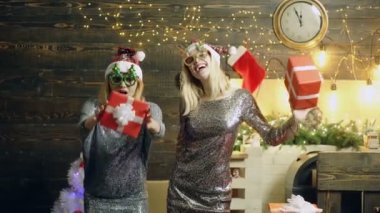 Noel kutlayan iki mutlu Noel Baba kızı ve evde yeni yıl hediyesi. Moda tarzı bir portre. İki kız arkadaşın ellerinde hediyeler, komik suratlar.
