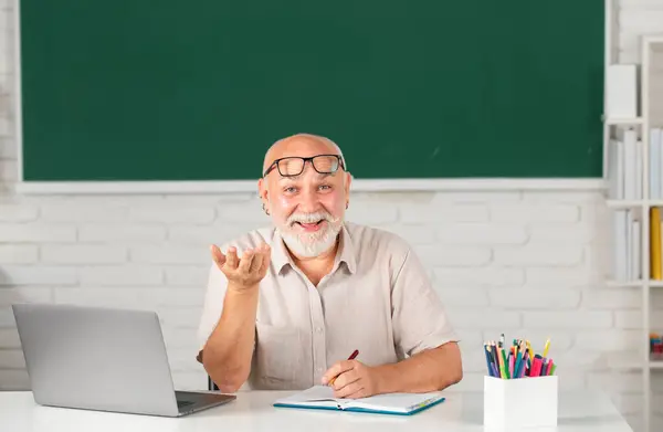 Man mature teacher. Portrait of happy senior tutor in glasses with book on the blackboard in class at high school or collage. Old student study in university. Adult teaching