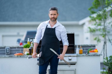 Man at a barbecue grill. Male cook preparing barbecue outdoors. Bbq meat, grill for picnic. Roasted beef. Attractive man preparing barbeque in the house yard. Barbecue and grill