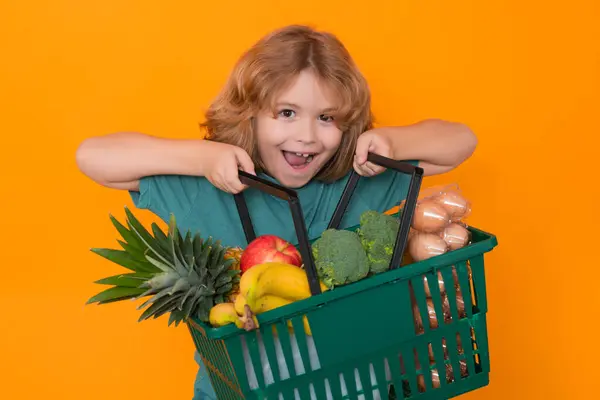 Grocery shop. Excited Child grocery cart, isolated studio yellow background with copy space. Little shopper. Kid holding shopping basket. Fresh organic, vegetables and fruits in grocery food store.