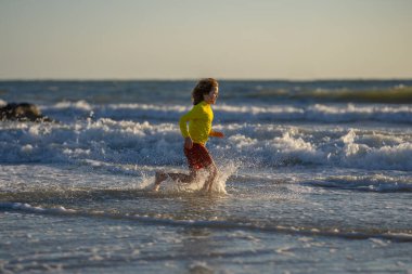 Gün doğumunda sahilde koşan küçük çocuk. Deniz sahilinde koşan mutlu çocuk. Yaz günü okyanus kenarında koşan sevimli bir çocuk. Çocuklar tropikal plajda koşuyor. Çocuk su sıçratarak oynuyor.