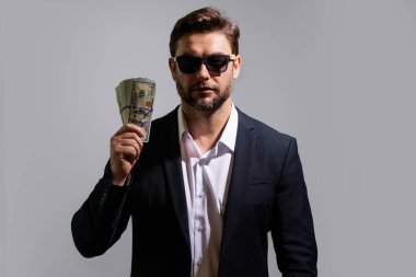 Nakit parayı dolar banknotlarıyla gösteren bir adam. Gri stüdyo arka planında izole edilmiş bir iş adamı portresi. Başarılı kazanan ya başarıyı ya da zaferi para faturalarıyla kutlar.