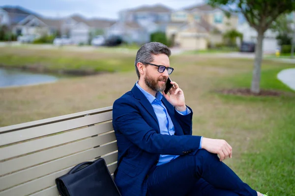 Business man in suit sitting on bench. Portrait of business man on bench in park outdoors. Thinking about business. Thoughtful businessman, freelancer work on laptop on american neighborhood. Free