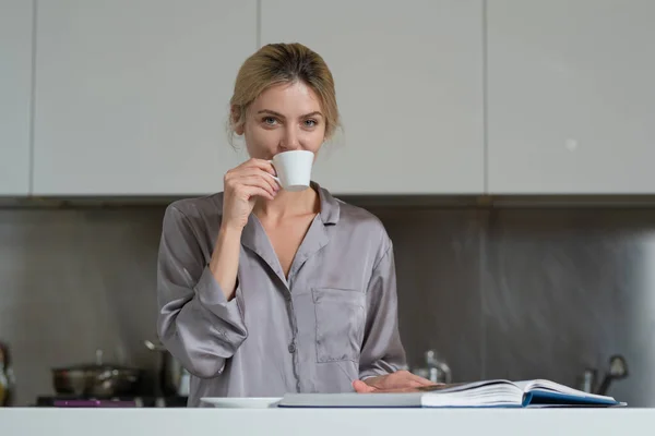 Sexy woman sitting in the kitchen in the morning and drinking coffee. Sensual housewife reading book or magazine