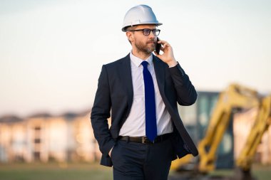 İnşaat mühendisi inşaat alanında çalışıyor. Olgun mühendis. İnşaat alanında takım elbiseli ve kasklı bir adam var. Orta yaşlı inşaat mühendisi kazıcının yanında duruyor.