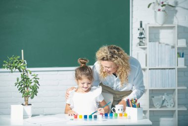 Anne ve kızı birlikte resim yapıyorlar. Öğretmen kız öğrenciye yardım ediyor.