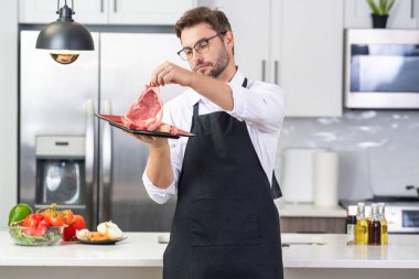 İspanyol 40 'lı yaşlardaki şef üniformalı adam mutfakta et tutuyor. Et pişiriyorum. Etli restoran menüsü. Yemek pişirmek, et reklamı yapmak. Çiğ biftek.