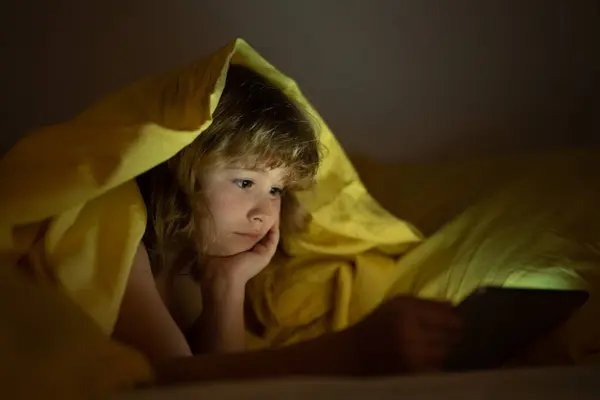 Kid alone at home. Kid watching tablet before sleeping. Child reads the e-book. Little boy looks at the screen of the tablet at hight. Kid boy playing tablet lying on a bed under blanket