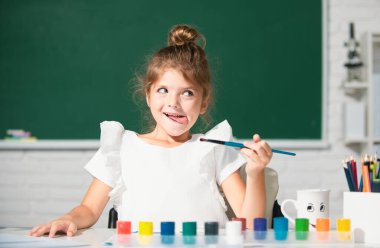 Çocuk kız sınıfta resim çiziyor, masada oturuyor, okulun karatahta arka planında eğleniyor. Küçük komik bir ressam, resim çiziyor. Komik çocuk duyguları.