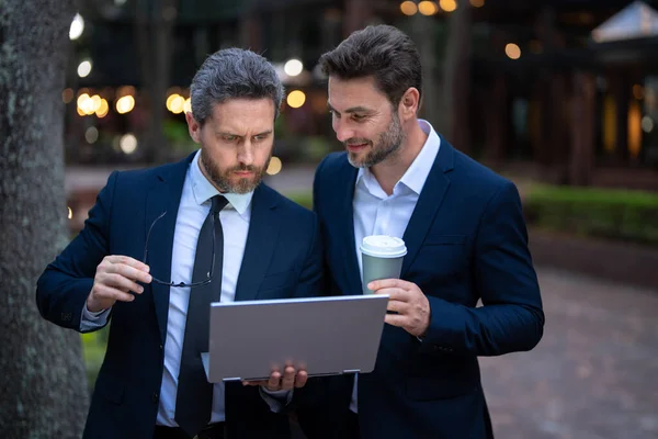 Two businessman using laptop outdoor. Freelance concept. Business men team using laptop outdoor. Two men talking in urban city center in business style working on laptop