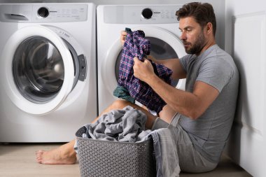 Çamaşır makinesinin yanında elbiseli bir adam. Yakışıklı adam çamaşır makinesinin önünde oturur. Çamaşır makinesini kirli çamaşırlarla dolduruyor. Adam elbiseleri temizliyor. Yalnız bir adam için ev işi. Ev çamaşırları