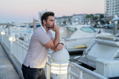 Yatın yanında hayal kuran ve düşünen zengin bir iş adamı. İyi giyimli, gözlüklü ve polo pozu veren bir adam. Erkek model. Şehirde şık bir adamın portresi. Açık hava moda portresi