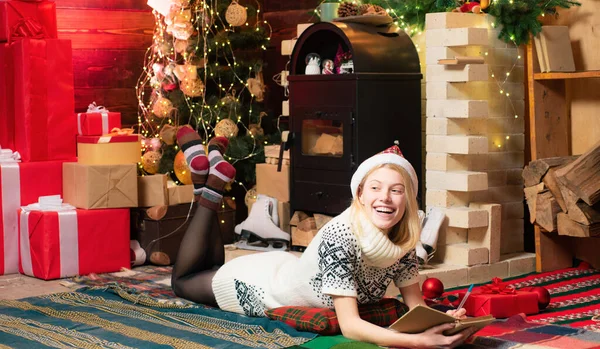 ニットセーターのドレスと赤い帽子を着た10代のブロンドの女の子は サンタにプレゼントのリストを書いています クリスマスの魔法の概念 家族と一緒に居心地の良い装飾された家で幸せな冬の休日 — ストック写真