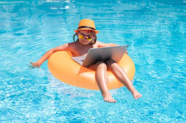 Küçük mutlu çocuk çevrimiçi bilgisayar bilgisayarı öğreniyor. Çocuk yüzme havuzunda bilgisayar kullanarak yüzen yüzükte rahatlıyor. Açık hava yaz işleri.